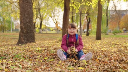 happy girl throws autumn yellow leaves up and laughs, kid is having fun playing in city nature park outdoors, little child with school backpack sits under leaf fall, first grader school yard having.