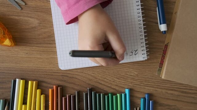 Top view of child doing math exercises in notebook with stationery on wooden table. Girl hand writing mathematical equations as school homework. Back to school, learning, education concept