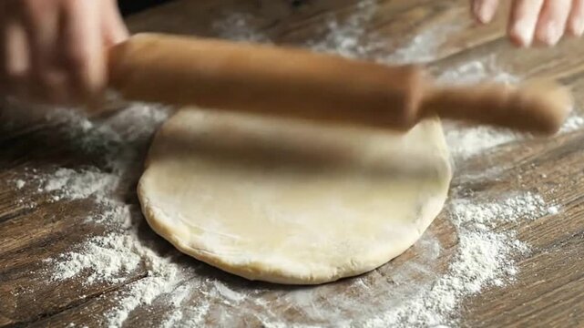 A person uses a rolling pin to flatten dough on a wooden table dusted with flour