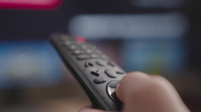 close-up of a caucasian hand with a TV remote switching channels, holding a TV remote control, flipping through channels and TV programs, looking for the desired movie in the cinema application.