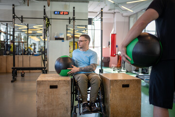 Disabled man in wheelchair do controlled exercises with weighted therapy ball, lifting stabilizing under trainer guidance. Determined athlete works on controlled ball lifts after severe spinal injury