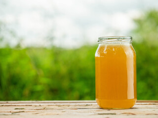Glass jar of golden bee honey is on a wooden table, green grass field and sky in the background. Fine organic nature product concept. Agriculture and food industry.