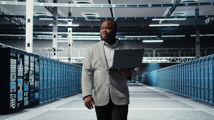 Smiling server room engineer performing maintenance on server racks using software on laptop. Happy data center IT professional upgrading blade server components using notebook, camera A