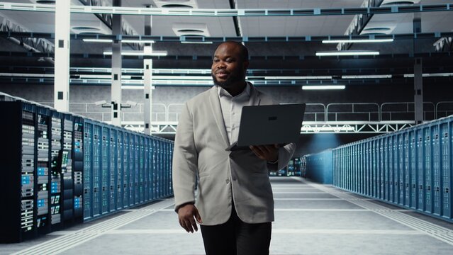 Smiling server room engineer performing maintenance on server racks using software on laptop. Happy data center IT professional upgrading blade server components using notebook, camera A