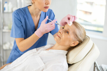 Obraz premium Beautiful elder woman receiving filler injection in interbrow zone at beauty clinic. Plastic surgery.