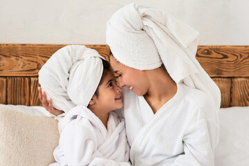 Sweet little girl having beauty day with her mother at home or at spa hotel, wearing white bathrobes, wrapping towels all over their heads, sitting on bed and cuddling, enjoying time together