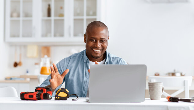 Happy african american handyman recording video about repairs and construction tools, looking at laptop webcam and sitting in kitchen interior with equipment on table, empty space