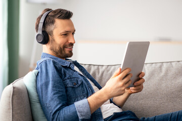 Joyful middle-aged bearded man laying on couch in living room, usind wireless headset and digital tablet, listening to music or watching videos online, using mobile apps, copy space