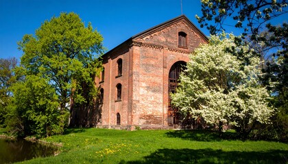 Fototapeta premium A brick building stands proudly beside a tranquil waterway, its facade adorned with many windows and detailed architecture, set against a vibrant spring day.
