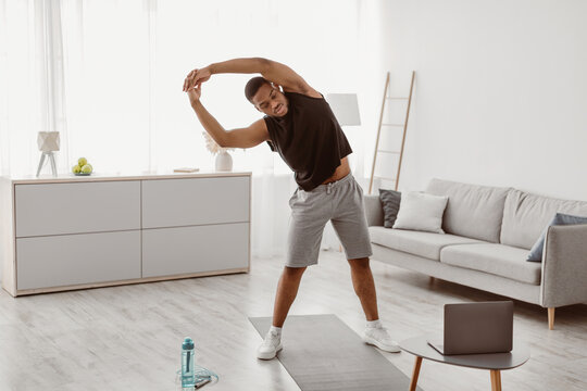 Online Male Fitness. Black Guy Exercising Near Laptop Doing Side Bend Stretching Standing In Living Room At Home. Workout Routine, Sporty Lifestyle Concept. Selective Focus - Powered by Adobe