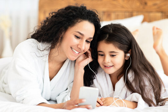 Pretty fair-haired young mother and little daughter in bathrobes laying on bed and watching videos on smartphone and smiling, using earphones, cozy bedroom interior, closeup portrait
