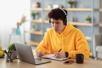 Portrait of focused asian man in headset and bright yellow hoodie sitting at desk, using pc writing in notebook, taking notes, watching tutorial, lecture or webinar, studying online at home