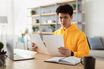Fototapeta premium Paperwork Concept. Portrait of focused asian manager working at home office, holding and reading paper or financial document, sitting at desk with laptop computer in living room, free copy space