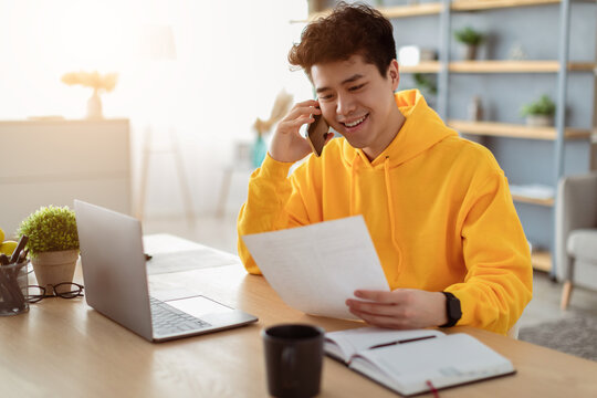 Corporate Communication. Portrait of smiling young asian male manager working remotely, holding paper and reading financial document, using laptop and talking on mobile phone, sitting at desk