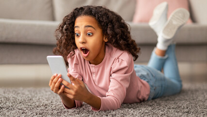 Wow. Emotional black girl reading unexpected message on smartphone, resting at home and lying on carpet. Overjoyed teen with open mouth holding cellphone and receiving great news