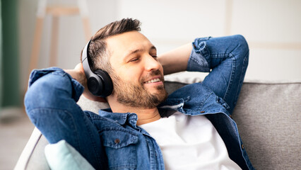 Time to relax. Joyful middle-aged bearded man in wireless headphones listening to music, copy space, closeup. Happy handsome man in headset with eyes closed relaxing on couch in living room