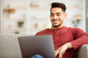 Closeup of happy middle-eastern man freelancer working from home, sitting on couch in living room and typing on notebook keyboard, chatting with clients or sending emails, copy space