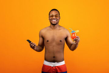 Shirtless black guy posing with smartphone and tasty tropical cocktail on yellow studio background. Funky young man holding mobile device and alcohol-free beverage, having summer vacation