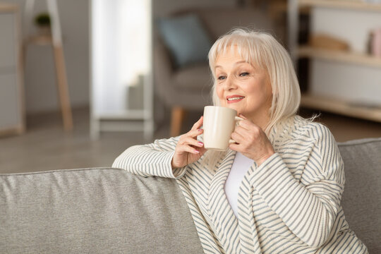 Weekend Leisure. Portrait of smiling mature woman drinking hot coffee, holding cup and sitting on the couch in living room. Happy senior lady enjoying good morning, blurred background, selective focus - Powered by Adobe
