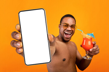 Excited black guy with naked torso drinking fresh fruit cocktail and taking selfie on yellow studio background. Emotional shirtless man enjoying refreshing tropical beverage on beach