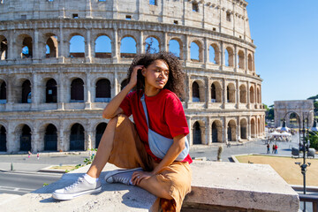 A stylish young woman in a chic outfit is sitting gracefully in front of the Colosseum, enjoying a sunny day while exploring Romes iconic historical site during her delightful adventure