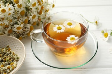 Aromatic tea in glass cup and chamomile flowers on white wooden table, closeup