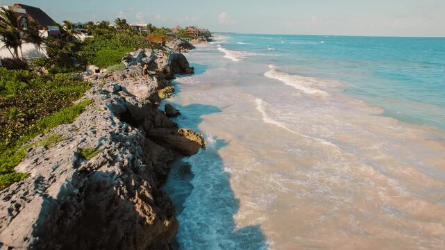 Woman stands on rocky cliff overlooking ocean waves at sunset with Tulum town in Mexico