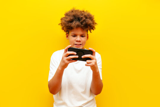 cheerful african american boy in white t-shirt playing video games on smartphone over yellow isolated background and smiling, curly schoolboy using phone for mobile games