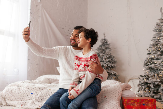 African American father and son in Christmas clothes taking selfie on smartphone at home, man and child celebrating Christmas and chatting on mobile online