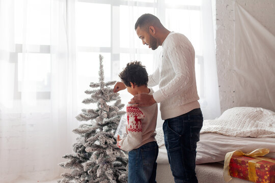 African American father and son in Christmas clothes decorating tree together at home, man helping child and celebrating Christmas together - Powered by Adobe