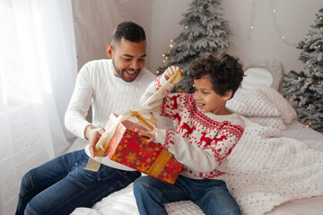 African American father giving gift to son for new year at home, boy opens gift box for christmas and is happy and surprised, family celebrate christmas in new year clothes