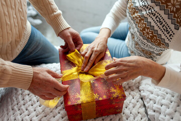 couple of elderly seniors in sweaters packing new year gift in red box, woman tying bow with gift ribbon on christmas, close up