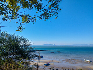 Scenic coastal view with calm blue sea, sandy shore, and small fishing boats under a clear blue sky, framed by tree branches