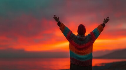 Fototapeta premium Person with arms raised in colorful sweater at vibrant sunset over water