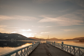 Obraz premium Scenic view of Worthersee lake from a wooden pier in Klagenfurt, Austria, with sunrise light reflecting on calm water and surrounding Carinthian mountains.