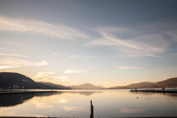 Wide view across Lake Worthersee at dusk in Klagenfurth, Austria. Calm water reflects mountains, sky and piers, with gentle golden light creating a peaceful winter lakeside landscape in Carinthia.