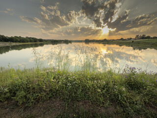Panorama of Markovacko Jezero lake near Mladenovac, Serbia. Calm lake water reflects clouds and sunbeams at golden hour, framed by grasses and wildflowers along the shore, a peaceful rural landscape.