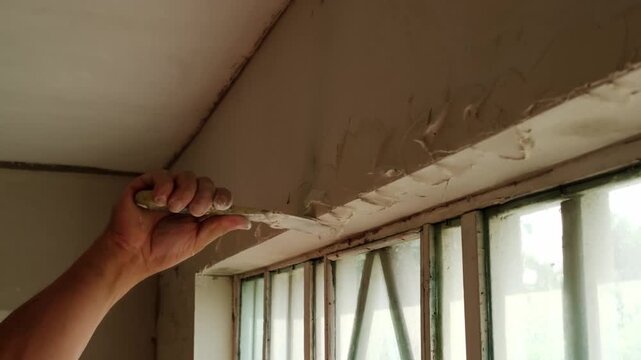 Plasterer holding putty knife smoothing wet plaster mix over window slope during indoor wall repair works.