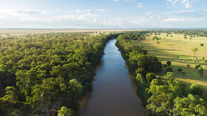 An aerial view of a wide, brown river flowing through a lush, green forest on one bank and open grasslands on the other, under a partly cloudy sky.