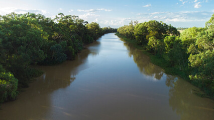 An aerial view of a wide, brown river flowing through a lush, green forest on one bank and open grasslands on the other, under a partly cloudy sky.