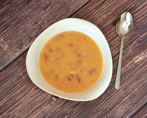 A deep bowl of delicious pea soup with smoked meat on a wooden table, a spoon nearby.