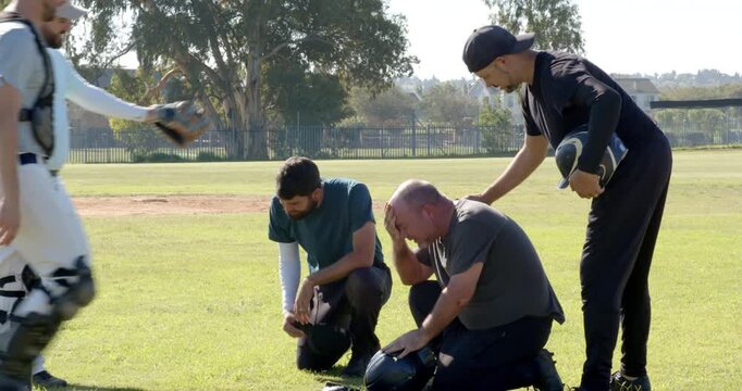 Walking across baseball field carrying helmets, gloves, male players helping injured teammate stand