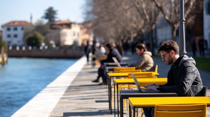 People working on laptops at yellow tables along a scenic riverside promenade on a sunny day