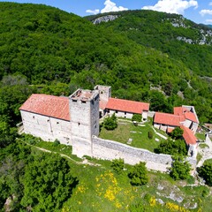 Aerial view of a medieval stone castle with red tile roofs, nestled within a verdant landscape of hills and trees