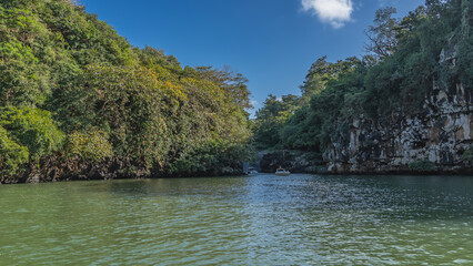 Naklejka premium A calm and beautiful tropical river. There is green vegetation on the steep banks. Tourist boats and a waterfall in the distance. Blue sky, clouds. Mauritius. Grand River South East waterfall
