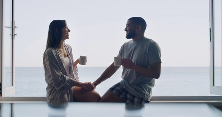 Ocean, window and couple with coffee, smile and bonding together with romance, talking and relationship. Home, seaside and man with woman, story and espresso with break, conversation or holding hands