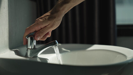 Close-up of male hand turning on off modern chrome faucet in bathroom. Wasting natural resources concept