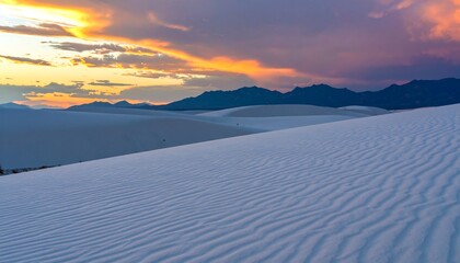 Undulating white sand dunes stretch towards a dramatic sunset, mountains silhouetted on the horizon