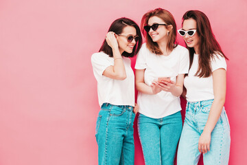 Three young beautiful smiling hipster female in trendy summer t-shirt and jeans clothes. Sexy carefree women posing near pink wall in studio.Positive models looking at smartphone screen. Using apps