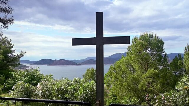 Slow motion zoom out footage shows a simple iron Christian  cross placed at the iron fence of a church. A lake and mountains in the background.